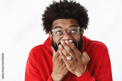 Waist-up shot of delighted happy african american male bearded friend in glasses chuckling, giggling with hand covering mouth looking up amused awaiting for prank come in action, joking over friend photo
