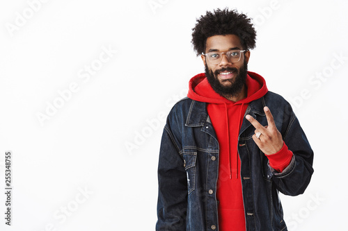 Portrait of satisfied and friendly good-looking african guy in glasses with beard posing in denim jacket over hoodie smiling and showing number two as making order or explaining on fingers