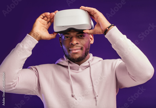 African handsome man dressed in white sports hoodie, taken off VR headset from his head, being impressed with virtual effects, smiling happily over violet purple background