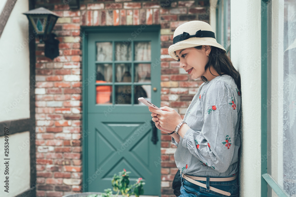 Beautiful teenager student girl using smart phone leaning against plain red stone texture brick wall networking and smiling outdoors. Adolescent technology lifestyle. vintage asian woman happy stand