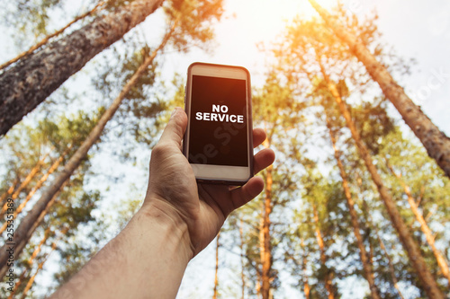 Male hand is holding a phone with sign no service  above his head against the background of trees in a pine forest. Concept of no service, wifi, internet in the forest or a place far from the city photo