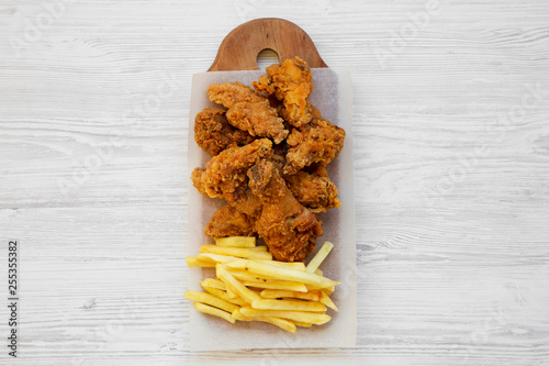 Tasty fastfood: fried chicken legs, spicy wings, French fries and chicken tenders on rustic wooden board over white wooden surface, top view. Flat lay, overhead, from above. photo
