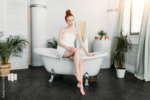 Gorgeous long-legged woman of 20 y.o. with ginger hair tied in knot on head, leaning on the edge of bathtub and touching her depilated legs. Hair removal or waxing removal beauty concept