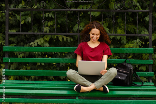 Image of cheerful happy young woman sitting on a bench in park and using her laptop, isolated outdoor on open air. Horizntal view. photo