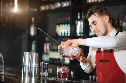 Smiling friendly looking barman wearing the bow tie, white shirt and red apron makes cocktail at bar counter at restaurant