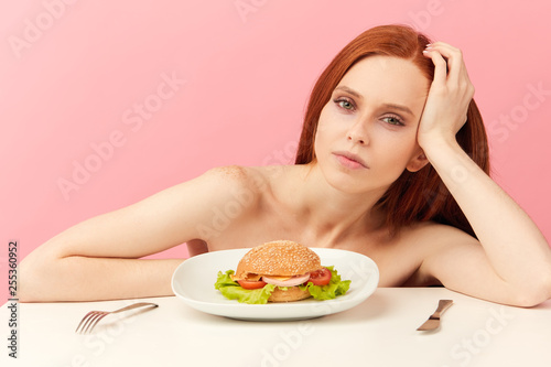 Diet failure of emaciated skinny redhead woman suffering from anarexia with greedy bugged eyes being ready to eat fast food hamberger. Unhealthy lifestyle bulimia concept. Junk meal leads