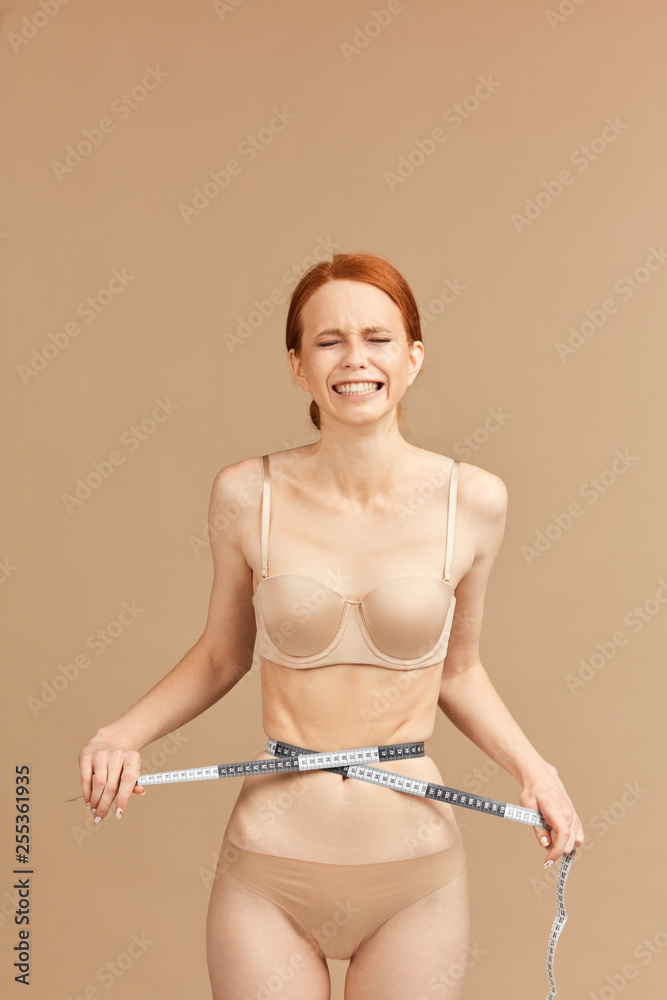 Skinny suffering female in nude underwear tied her waist with measuring  tape posing over beige background in studio. Anorexia and eating disorders  Photos
