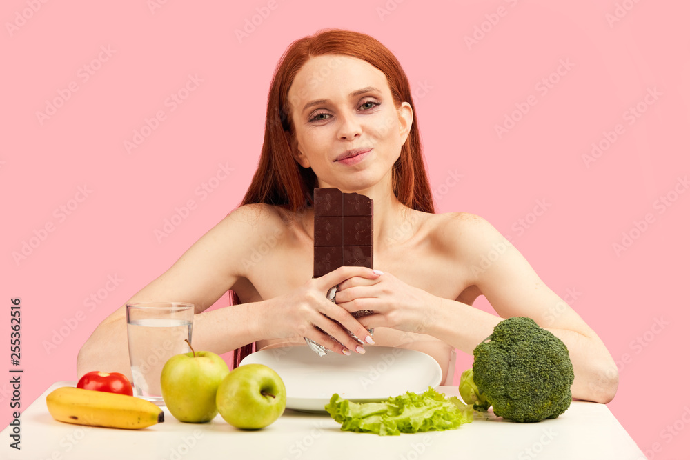 Young woman with skinny complexion and unhealthy pale skin, is bored of restrictions feeling happy for eating sweet chocolate bar instead of healthy food. Diet failure or eating disorders. Young woman with skinny complexion and unhealthy pale skin, is bored of restrictions feeling happy for eating sweet chocolate bar instead of healthy food. Diet failure or eating disorders.