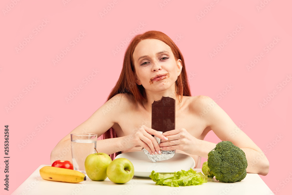 Happy young skinny woman exhausted by severe diets, holding chocolate bar, posing isolated on pink. Diet failure of emaciated redhead woman is eating unhealthy forbidden food with enormous pleasure. Happy young skinny woman exhausted by severe diets, holding chocolate bar, posing isolated on pink. Diet failure of emaciated redhead woman is eating unhealthy forbidden food with enormous pleasure.