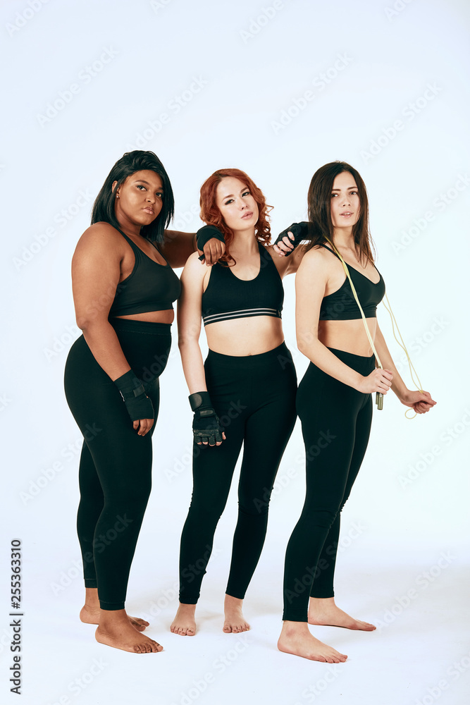 Fototapeta premium African happy overweight woman in company of two caucasian slim girlfriends, dressed in sportive clothes . Weight and sport. The reason why weight is growing during sports training.