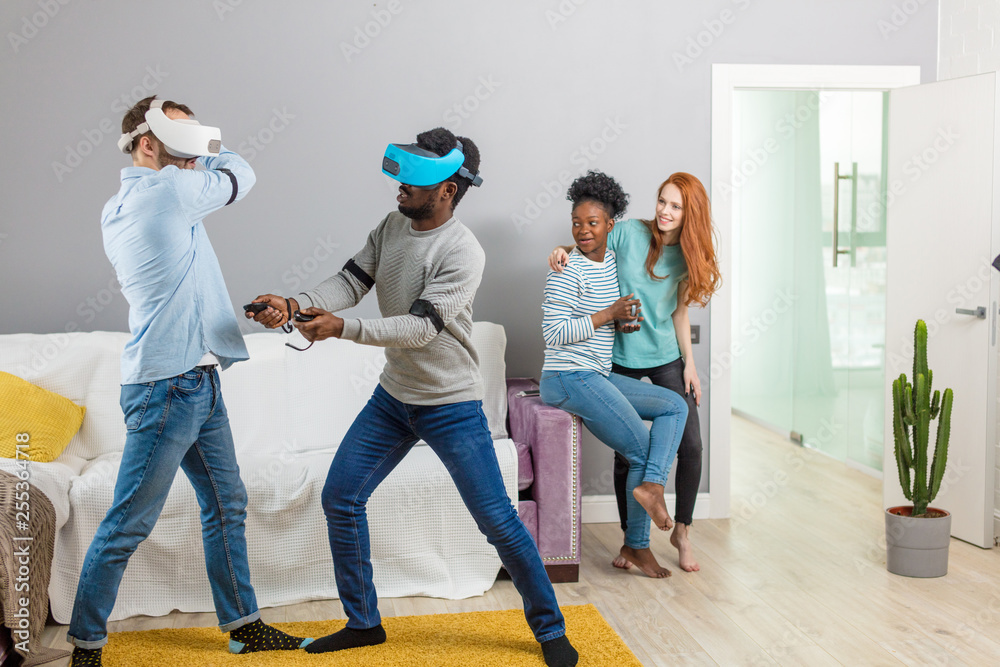 International diverse students resting together with glasses of virtual reality an home, two young interracial men entertaining girls International diverse students resting together with glasses of virtual reality an home, two young interracial men entertaining girls