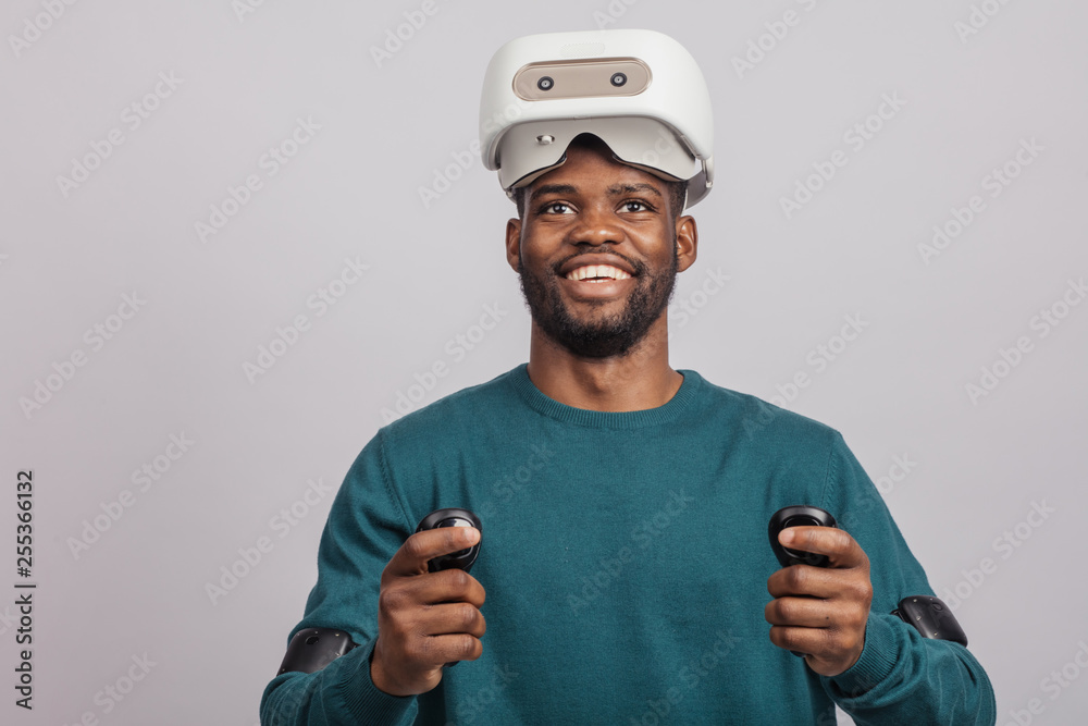 Young dark skinned guy getting excited after watching 3 D film using virtual reality headset, looking at camera isolated in green wear over grey background. Young dark skinned guy getting excited after watching 3 D film using virtual reality headset, looking at camera isolated in green wear over grey background.