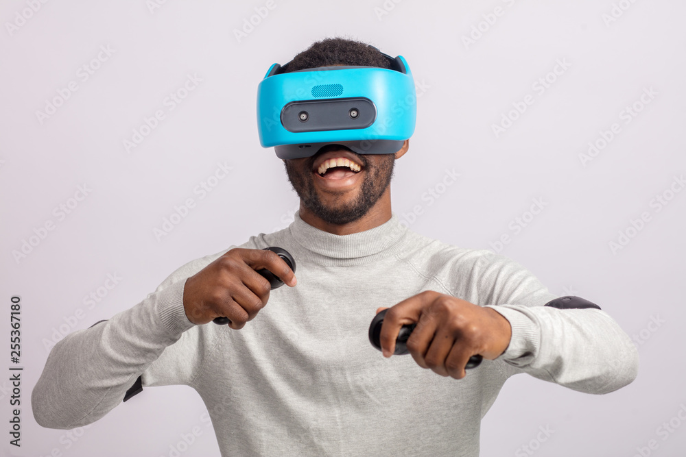 Naklejka premium African american guy in casual wear plays racing game in VR headset, holding hands with controllers raised as if he is driving car on high speed in virtual reality.