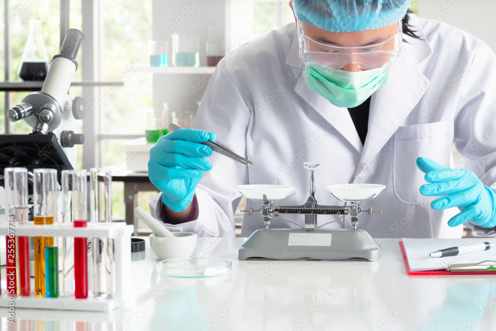 An asian chemist or scientist researcher using mechanical scale for gathering and measuring specimen at laboratory. The young man wearing safety glasses in experiment. Pharmaceutical research concept An asian chemist or scientist researcher using mechanical scale for gathering and measuring specimen at laboratory. The young man wearing safety glasses in experiment. Pharmaceutical research concept