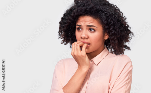 Anxious fearfull African American young woman keeps hand near mouth, feels frightened and scared, wearing in beige shirt, standing against white studio wall. Facial expressions, emotions and feelings