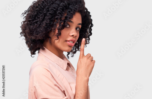 Confidential African American young woman dressed in pink shirt holding index finger on lips, asking to keep silence over white wall. Beautiful dark skinned female looks to the camera asks to be quiet