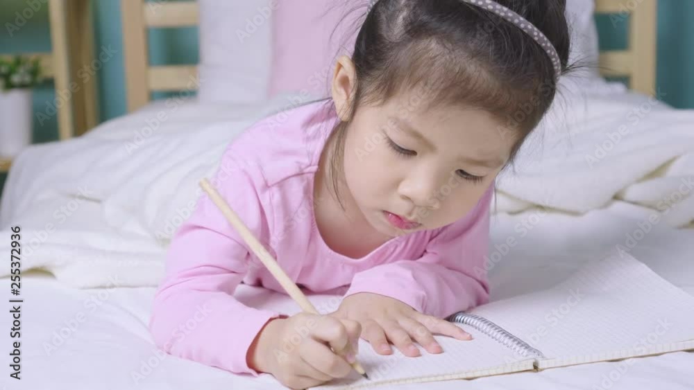 Happy child little asian girl writing in a book on the bed in the bedroom, Educational concept for homeschool Happy child little asian girl writing in a book on the bed in the bedroom, Educational concept for homeschool