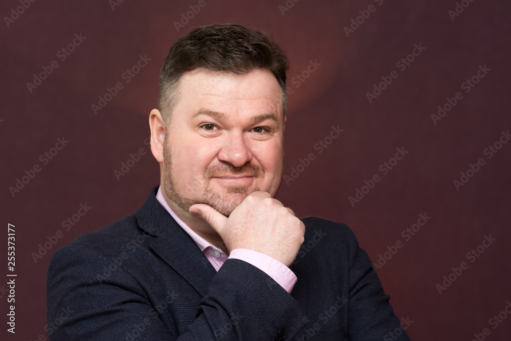 A middle-aged man, a large portrait. A man with a beard in a suit on a dark background raised his hand to his chin. A middle-aged man, a large portrait. A man with a beard in a suit on a dark background raised his hand to his chin.