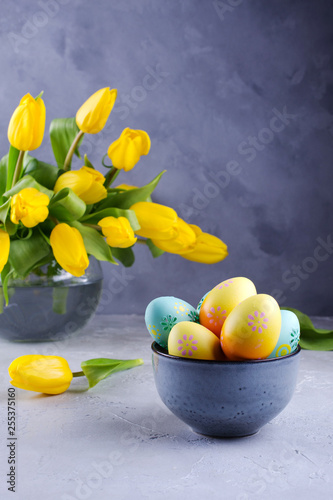 Bowl with colorful Easter eggs; spring easter decoration on gray table with bouquet of yellow tulip flowers in glass vase; Easter interior decoration