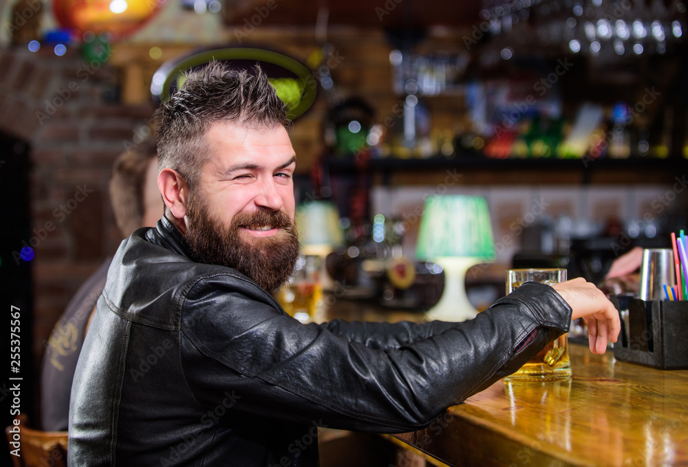 Hipster relaxing at bar with beer. Brutal hipster bearded man sit at bar counter drink beer. Order alcohol drink. Bar is relaxing place have drink and relax. Man with beard spend leisure in dark bar