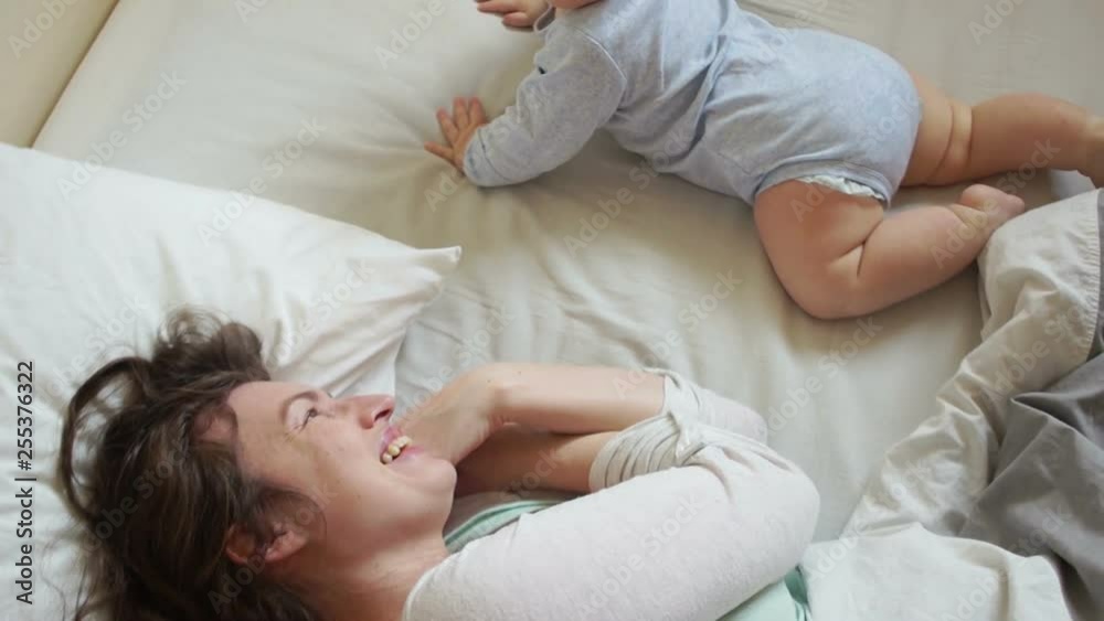 Caring mother and baby are playing in bed in the morning. Baby rolls over and crawls, mother returns him back, baby laughs Caring mother and baby are playing in bed in the morning. Baby rolls over and crawls, mother returns him back, baby laughs