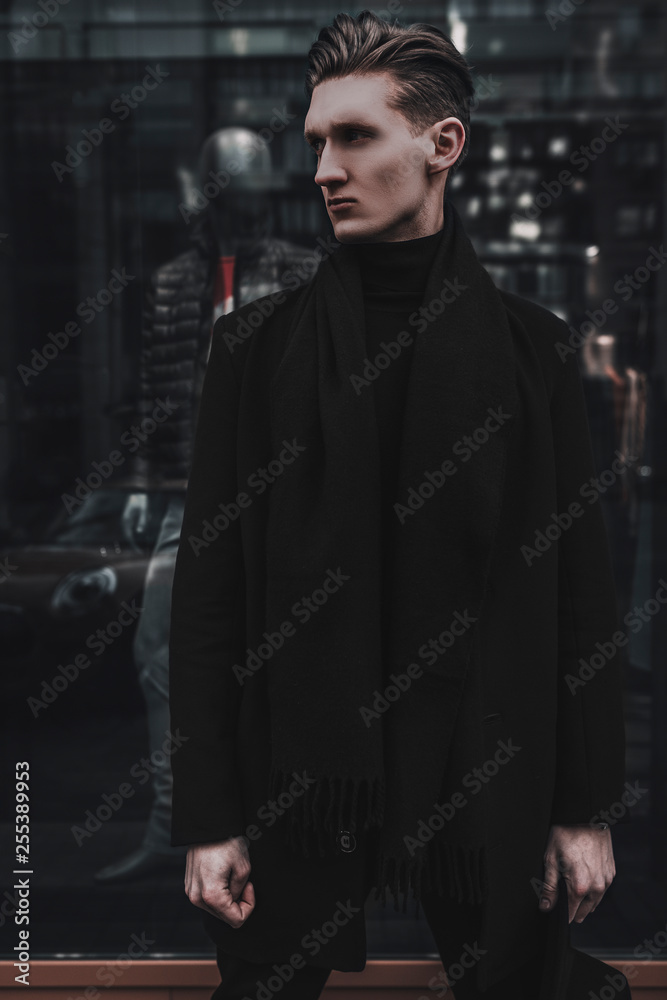 Stylish fashion young man model posing near shop window on city street. Urban male model test. Wearing in black coat and scarf.. Stylish fashion young man model posing near shop window on city street. Urban male model test. Wearing in black coat and scarf..
