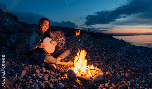 Obraz na plátně cheerful percussionist girl playing djembe sitting on beach by the fire at sunset