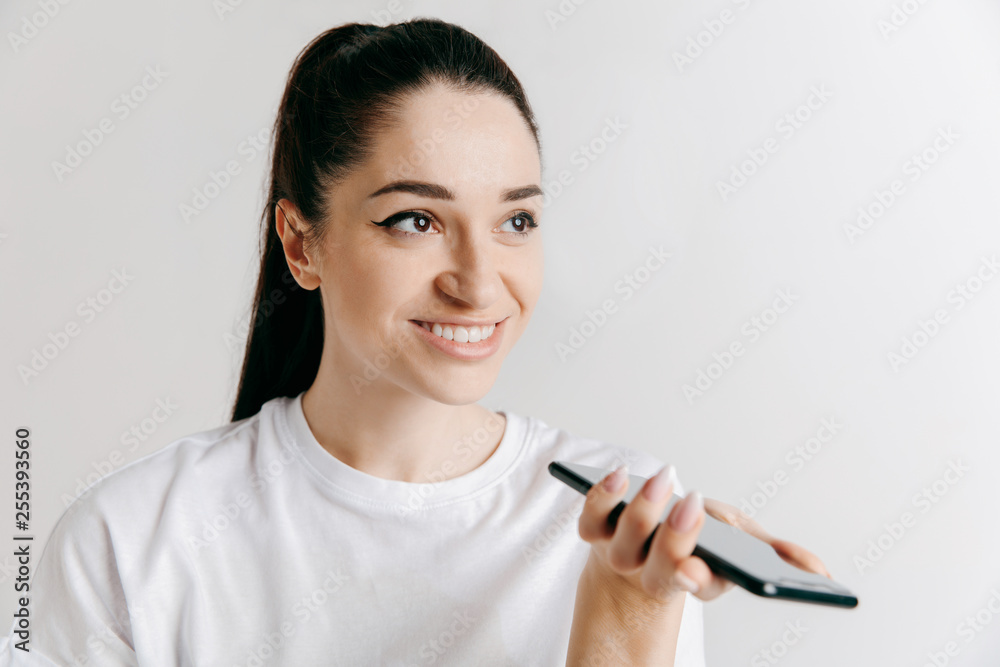 Indoor portrait of attractive young woman isolated on gray background, holding blank smartphone, using voice control, feeling happy and surprised. Human emotions, facial expression concept. Indoor portrait of attractive young woman isolated on gray background, holding blank smartphone, using voice control, feeling happy and surprised. Human emotions, facial expression concept.