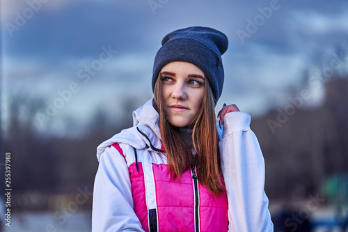 Portrait of a young beautiful girl with dark brown hair in a sports hat and jacket in the first rays of the morning rising sun in an autumn cold morning.