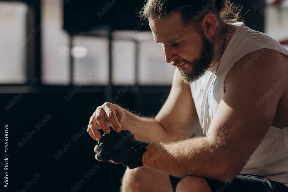 Boxer puts on hand wraps while sitting on the edge of a boxing ring in a boxing gym, camera rotates around