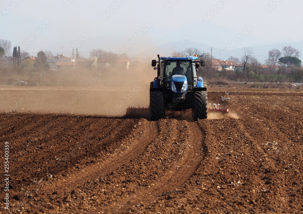Obraz premium Tractor plows the dry agricultural field raising dust, in a sunny morning of early springtime