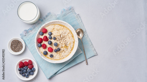 Oatmeal with berries, chia, maple syrup and glass of milk on blue light background. Top view, copy space. Long width banner. Healthy diet breakfast