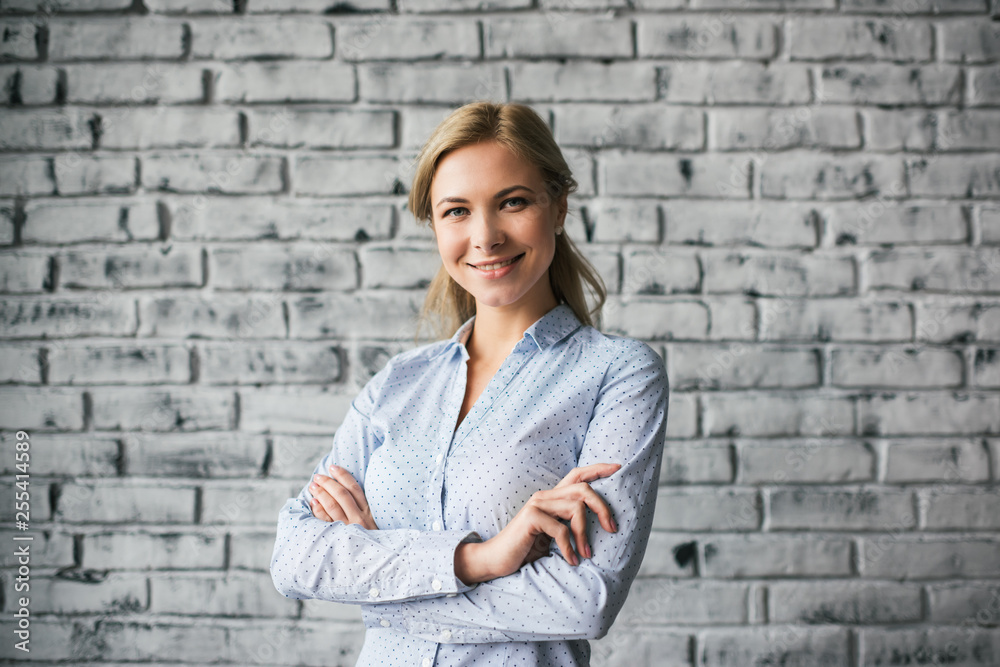  Happy business woman with arms crossed  posing for pictures in loft office isolated on background. Successful business concept. Portrait of young cheerful top manager standing near grey wall.  