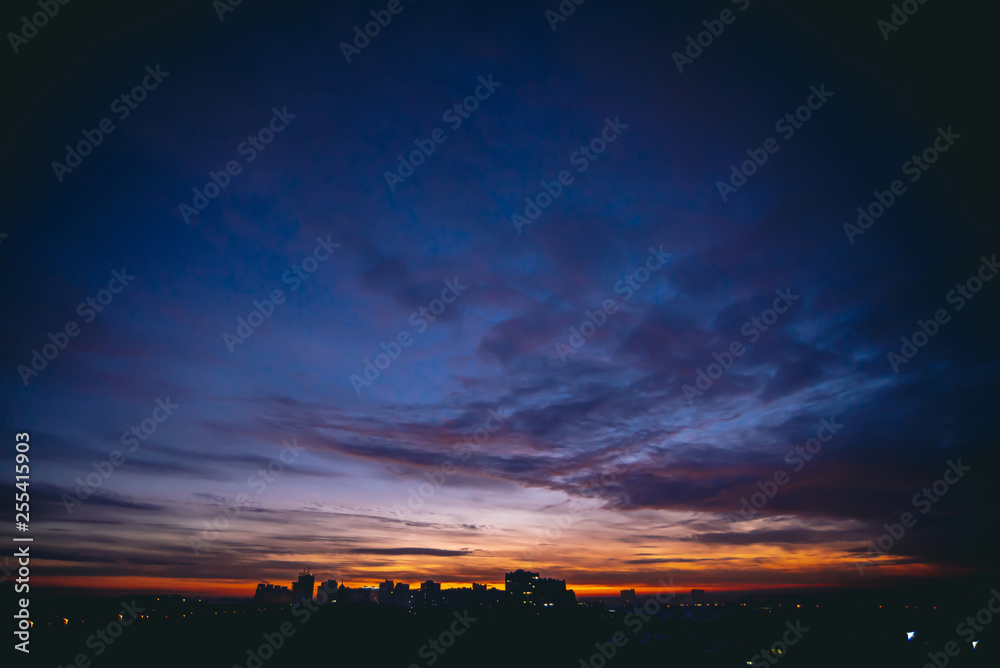 Cityscape with vivid warm dawn. Amazing dramatic blue violet cloudy sky above dark silhouettes of city buildings. Orange sunlight. Atmospheric background of sunrise in overcast weather. Copy space. Cityscape with vivid warm dawn. Amazing dramatic blue violet cloudy sky above dark silhouettes of city buildings. Orange sunlight. Atmospheric background of sunrise in overcast weather. Copy space.