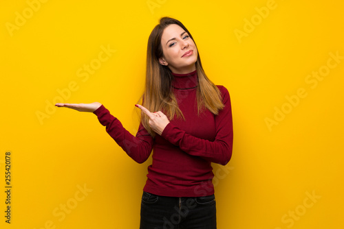Woman with turtleneck over yellow wall holding copyspace imaginary on the palm to insert an ad © luismolinero