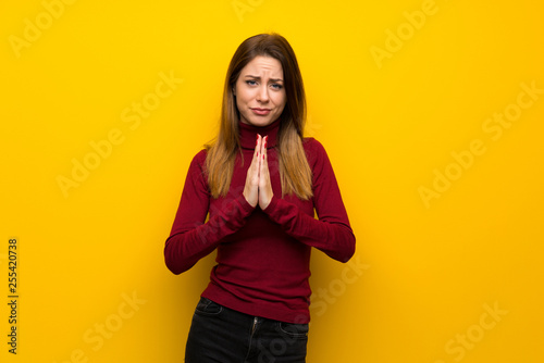 Woman with turtleneck over yellow wall keeps palm together. Person asks for something © luismolinero