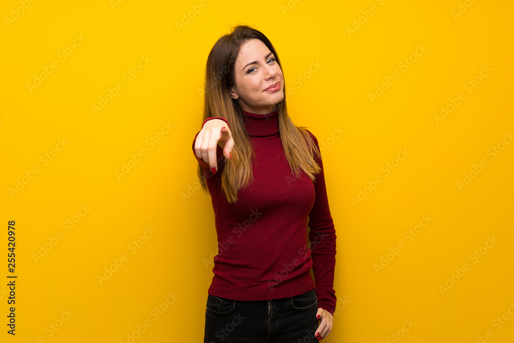 Woman with turtleneck over yellow wall points finger at you with a confident expression