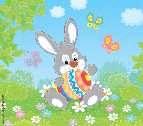 Little Easter Bunny sitting with a colorfully decorated big egg among flowers and flittering butterflies on a green lawn on a sunny spring day, vector illustration in a cartoon style