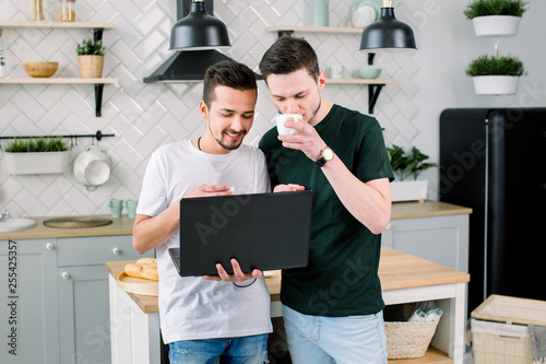 Two happy friends having fun while using laptop together. Happy smiling gay couple using laptop at the kitchen at home photo
