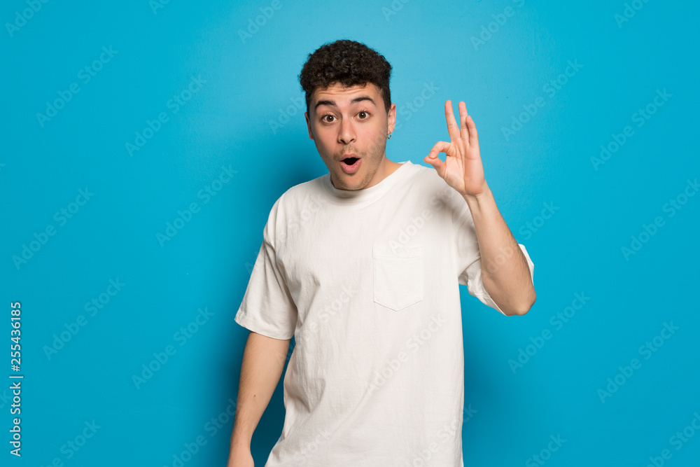 Young man over blue background surprised and showing ok sign Young man over blue background surprised and showing ok sign
