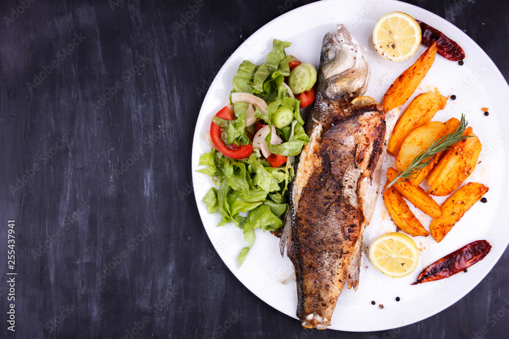 Fried sea bass with lemon, potatoes, salad and spices on a black wooden table