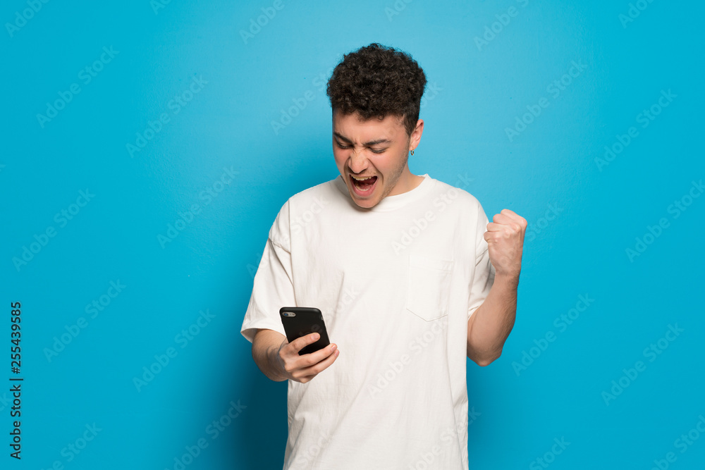 Young man over blue background celebrating a victory with a mobile Young man over blue background celebrating a victory with a mobile