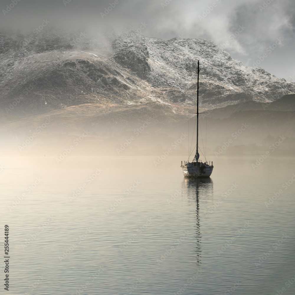 Fototapeta Stunning landscape image of sailing yacht sitting still in calm lake water with mountain looming in background during Autumn Fall sunrise