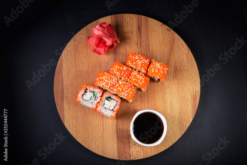 Sushi California Roll with crab meat, cucumber, masago, soy sauce and ginger on a round wooden cutting board, black background, top view