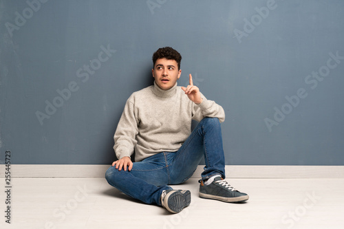 Young man sitting on the floor thinking an idea pointing the finger up