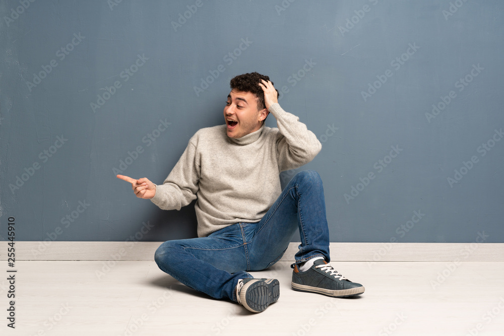 Young man sitting on the floor surprised and pointing finger to the side Young man sitting on the floor surprised and pointing finger to the side