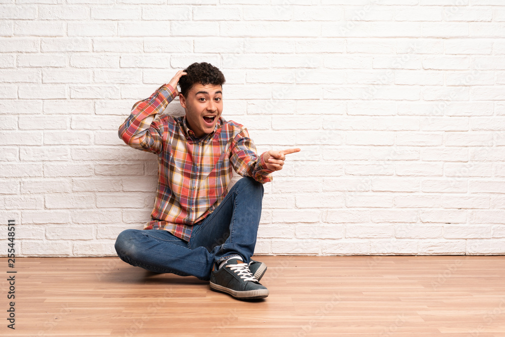 Young man sitting on the floor surprised and pointing finger to the side Young man sitting on the floor surprised and pointing finger to the side