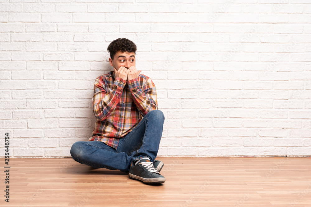 Young man sitting on the floor nervous and scared putting hands to mouth Young man sitting on the floor nervous and scared putting hands to mouth