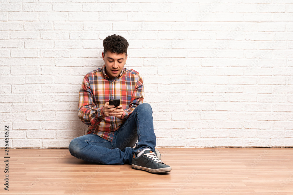 Young man sitting on the floor sending a message with the mobile Young man sitting on the floor sending a message with the mobile