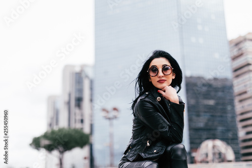 Casual style. Stylish woman in sunglasses sitting on the stone stairs. She dressed in leather jacket, skinny.
